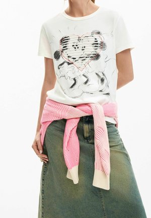 Femme portant un T-shirt blanc avec deux personnages de dessin animé à l’intérieur d’un cœur cousu, pull rose noué autour de la taille et jupe vert olive.
