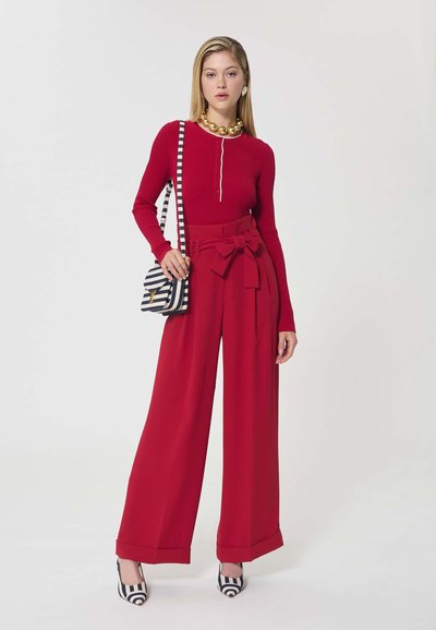 Femme portant un top rouge à manches longues et un pantalon large rouge avec un nœud, sac rayé sur l'épaule, grand collier en or, et chaussures à talons rayées.