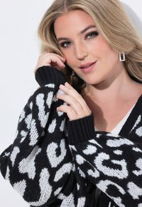 Svart och vitmönstrad stickad cardigan med ribbade ärmslut, med en avslappnad passform och mjuk textur, bärs över en vit topp.