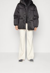 Černá péřová bunda se zipem a patentkami, texturovaná tkanina, kapuce a diamantové prošívání, spárovaná s bílými kalhotami s širokými nohavicemi.