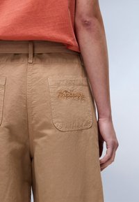 Beige cargobroeken hebben een kastanjekleurig geborduurd logo op de achterzak, met een relaxte pasvorm en een fijne katoenen stof.