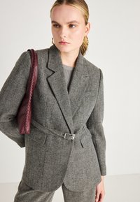 Sivi herringbone blazer s pojasom u struku, revere i dva prednja džepa. Uparen s burgundskom kožnom torbicom.