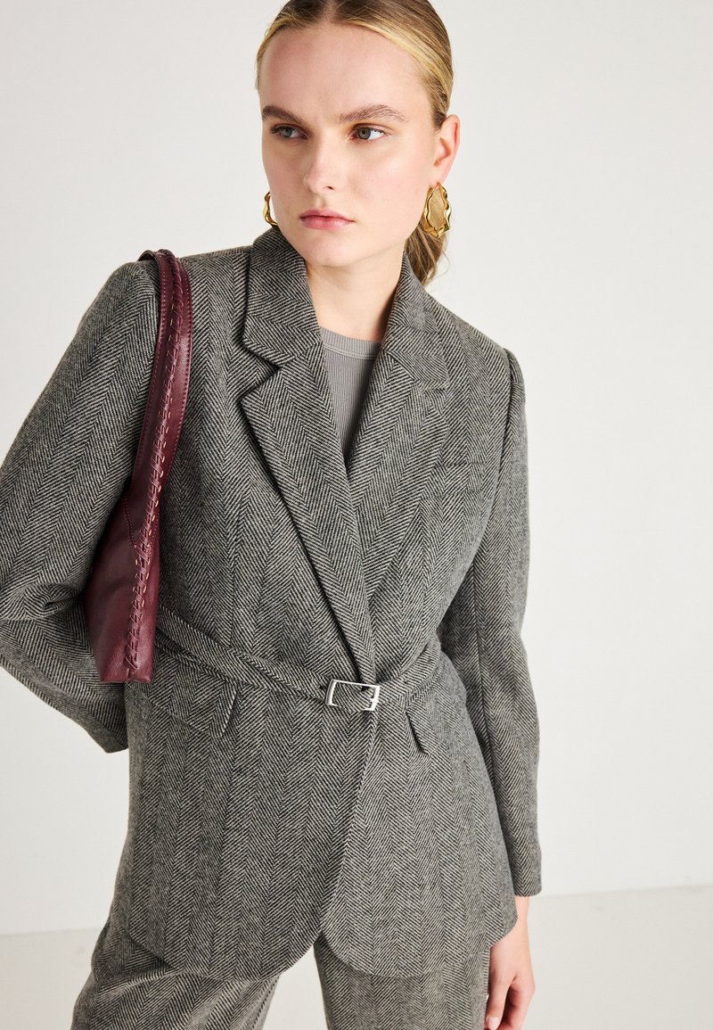 Sivi herringbone blazer s pojasom u struku, revere i dva prednja džepa. Uparen s burgundskom kožnom torbicom.