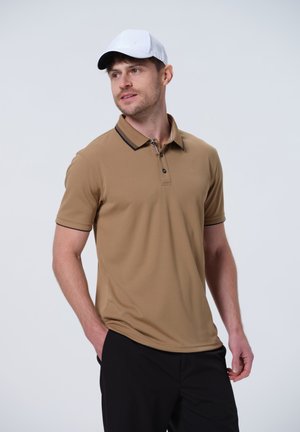 Camiseta polo de color marrón claro con mangas cortas, detalles en negro en el cuello, tapeta de dos botones y tejido que absorbe la humedad. Combinada con pantalones negros.