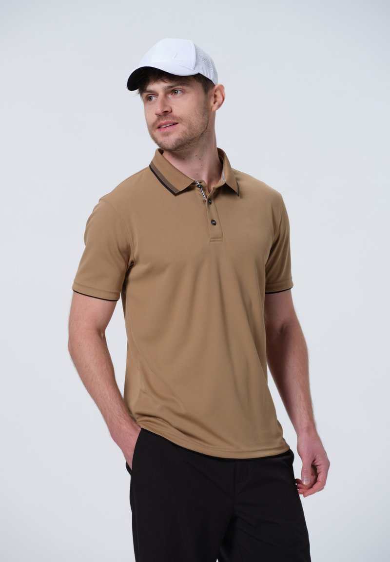 Camiseta polo de color marrón claro con mangas cortas, detalles en negro en el cuello, tapeta de dos botones y tejido que absorbe la humedad. Combinada con pantalones negros.