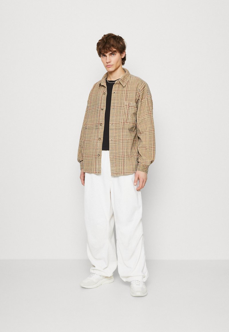 Brown checkered shirt jacket with a relaxed fit, paired with white wide-leg pants and light-colored sneakers. Simple background.