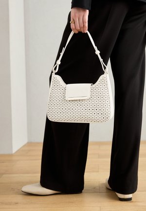Witte handtas met een geperforeerd ontwerp, gestructureerde vorm en een solide klepsluiting. Gekocht met zwarte broeken op een houten vloer.