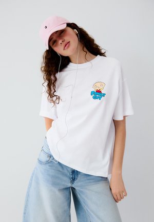 Giovane donna indossa un berretto rosa, ascolta gli auricolari e posa con una T-shirt bianca di Family Guy e jeans azzurri davanti a uno sfondo semplice.