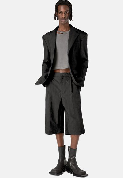 Blazer negro oversized sobre una camiseta gris cropped, combinado con pantalones cortos a medida y botas negras con cremallera. Tejido texturizado con un ajuste estructurado.