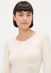 Haut long à manches raglan de couleur crème claire avec un col rond, mettant en avant une petite étiquette en tissu près du col et une texture lisse.