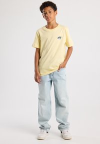 Lichtgele katoenen t-shirt met een klein blauw "SURF"-motief, gecombineerd met lichtblauwe baggy jeans en witte sneakers.