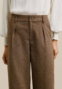 Pantalons en chevrons marron avec des plis, une fermeture à bouton et un tissu texturé. Associés à un chemisier blanc drapé délicatement.