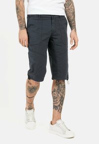 Marinefarbene Cargo-Shorts mit mehreren Taschen, knielang, aus leichtem Material. Kombiniert mit weißen Sneakers und tätowierten Beinen.