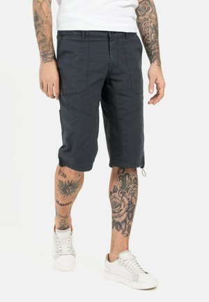 Marine cargo shorts med flere lommer, knæ-lange, lavet af letvægtsstoffer. Parret med hvide sneakers og viser tatoverede ben.