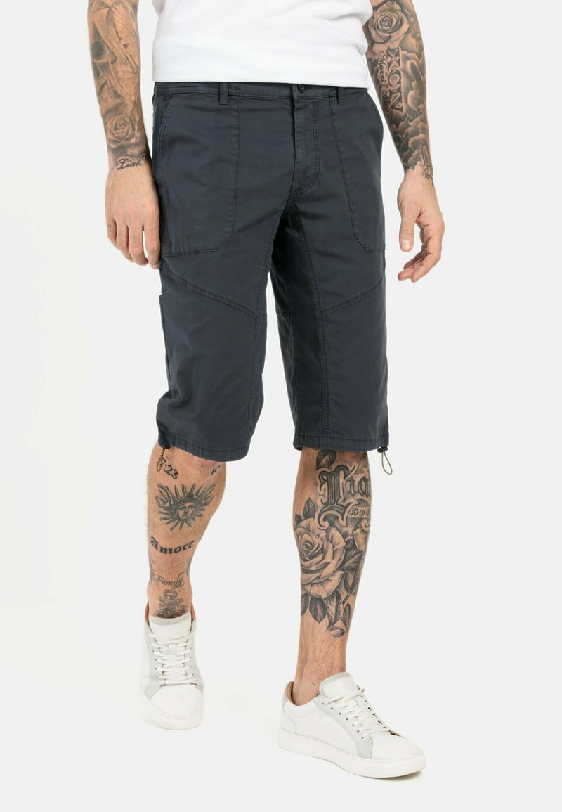 Marinefarbene Cargo-Shorts mit mehreren Taschen, knielang, aus leichtem Material. Kombiniert mit weißen Sneakers und tätowierten Beinen.