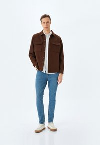 Chaqueta de lana marrón con dos bolsillos frontales, cierre de botones y cuello puntiagudo, combinada con jeans de mezclilla azules y zapatillas beige.