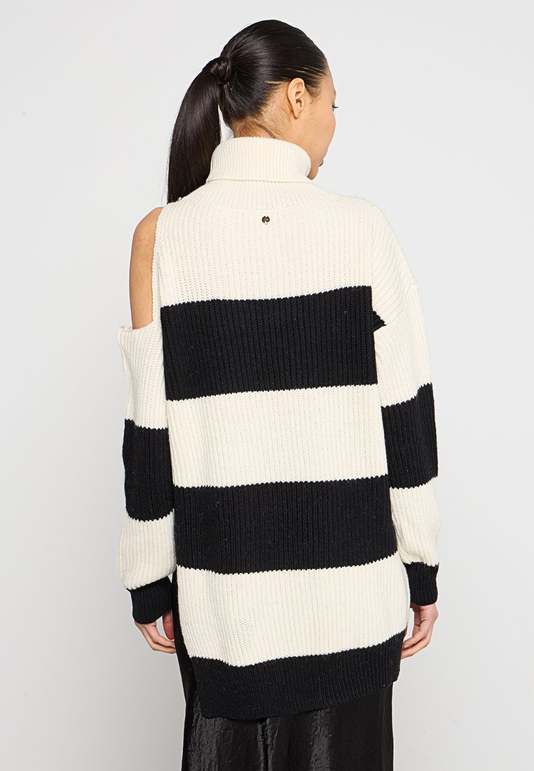 Vrouw met een zwarte paardenstaart die een lange, geribbelde zwart-wit horizontaal gestreepte trui met een schouderuitsnijding draagt, met haar rug naar ons gekeerd.