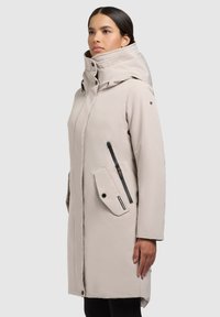 Vrouw in een lichtbeige lange gewatteerde winterjas met hoge kraag, capuchon en zwarte ritszakken, staande tegen een effen achtergrond.