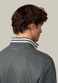Grijze poloshirt met een hoge kraag, met witte en zwarte gestreepte accenten. Logo "Hackett" in goud op de achterkant. Zachte textuur.