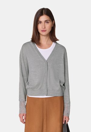 Hellgrauer Cardigan aus weichem Material, mit V-Ausschnitt, lockerer Passform und Spitzenakzenten an den Ärmeln. Kombiniert mit einem weißen Oberteil.