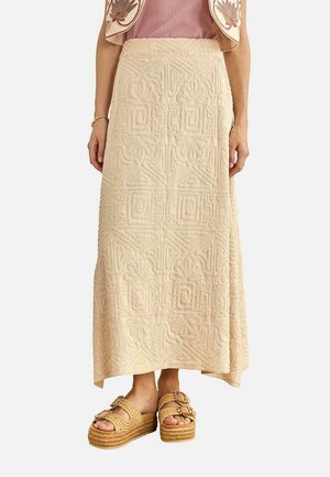 Mujer con falda maxi beige texturizada con patrones geométricos, combinada con sandalias de plataforma y un chaleco sin mangas bordado.