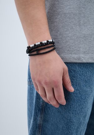 Melnas un baltas pērļu aproces sakrautas uz plaukstas, ar dažādām pērļu formām un tekstūrām, ar pinumiem no melnas ādas akcentiem.