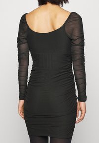 Svart klänning med långa genomskinliga ärmar, rynkad kropp och rundad rygg. Tillverkad av stretchigt material som framhäver en åtsittande silhuett.