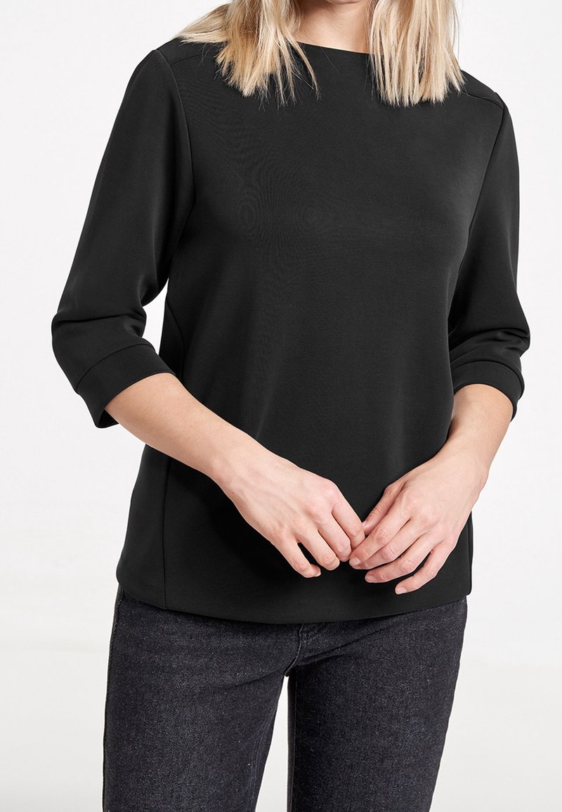 Schwarzes, langärmliges Oberteil mit rundem Halsausschnitt und umgeschlagenen Manschetten. Glattes Material mit lockerer Passform, getragen mit dunklen Jeans. Flache Nahtdetails.