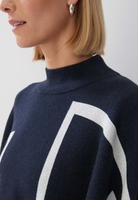 Dunkelblauer Strickpullover mit hohem Kragen und weißem geometrischem Muster. Weiche Textur und entspannter Schnitt. Sichtbarer silberner Ohrhänger.