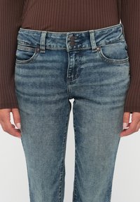 Mittelteil einer Person, die ausgewaschene blaue Jeans mit zwei Knöpfen und ein braunes, geripptes Langarmoberteil trägt, die Hände entspannt an den Seiten liegend.