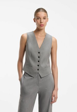 Femme portant un gilet sans manches gris à fines rayures avec des boutons noirs et un pantalon large assorti, debout avec une main dans la poche.