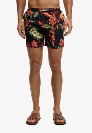 Mann trägt schwarze Badeshorts mit rotem und grünem tropischem Blumenmuster und braune Ledersandalen, steht vor weißem Hintergrund.