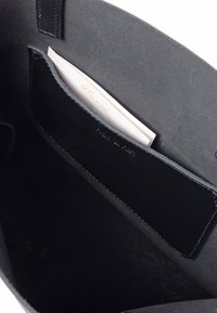 Wnętrze czarnej skórzanej torby typu tote, charakteryzujące się gładką fakturą, eleganckim designem oraz kieszenią z widocznym złożonym kawałkiem papieru w środku.