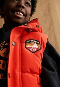 Doudoune orange vif avec un patch logo Superdry Everest, des accents noirs, des boutons pression et un tissu texturé.