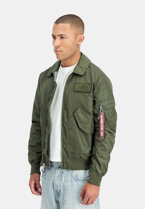 Man wearing olive green Alpha Industries bomber jacket with zip, flap pockets, and "Remove Before Flight" tag on sleeve, white shirt, light jeans.