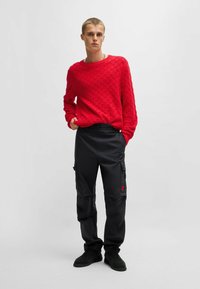 Maglione rosso lavorato a maglia con un motivo testurizzato, abbinato a pantaloni cargo neri con tasche laterali e un piccolo logo rosso. Calzature nere.