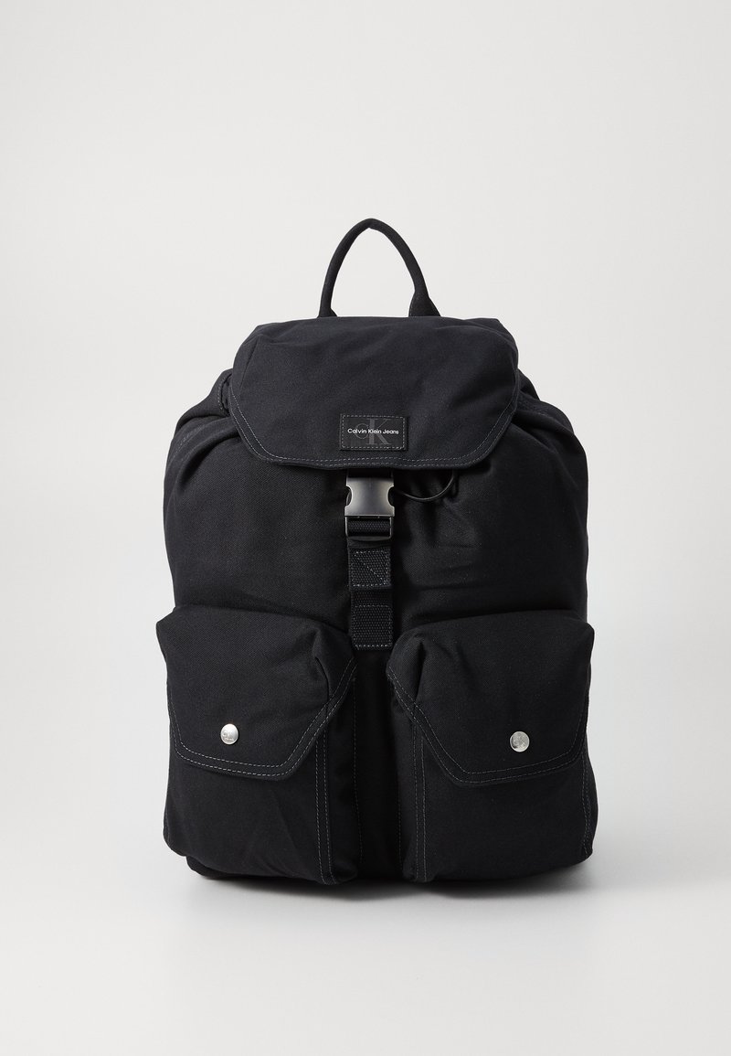 Black canvas backpack with a flap closure, silver hardware, two front pockets, and a top handle. Notable stitching detail emphasizes the design.