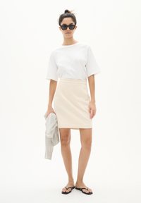 Haut blanc à manches courtes associé à une jupe beige jusqu'aux genoux, des tongs noires et tenant un vêtement gris. Design minimaliste et couleurs classiques.
