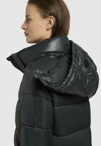 Puchová bunda v tmavošedej farbe, s prešívaným dizajnom, oversized kapucňou a prednými zipsovými vreckami s brandingom na golieri a zipsy.