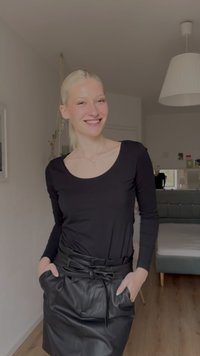 Mlada ženska, oblečena v črn dolg rokav in usnjeno krilo, se smehlja z rokami v žepih v svetli, sodobni spalnici.