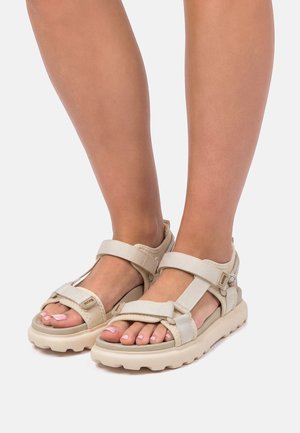 Sandali beige aperti con cinturini regolabili indossati su piedi con unghie dei piedi dipinte di rosa chiaro su uno sfondo neutro.