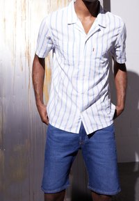 Camisa de manga corta abotonada en blanco con rayas azules, combinada con shorts de mezclilla, con un bolsillo delantero y corte relajado.