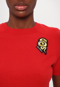 Rotes gestricktes Kurzarmshirt mit einem schwarz-goldenen gestickten Schlangenlogo und dem Text "MAX&CO" auf der linken Seite.