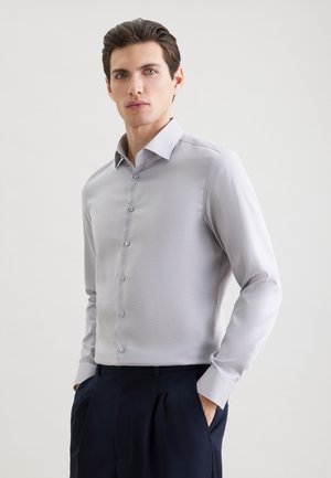 Młody mężczyzna o ciemnych włosach, ubrany w jasnoszarą, zapinaną na guziki koszulę i ciemne spodnie, stojący z rękami w kieszeniach na jednolitym tle.