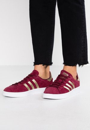 Stopy w bordowych zamszowych butach sportowych Adidas ze złotymi paskami i białą podeszwą, połączone z czarnymi spodniami z postrzępionym dołem.