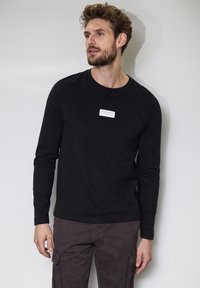 Black long-sleeve shirt with a crew neck and a small white label on the chest, paired with dark cargo pants. Smooth cotton texture.