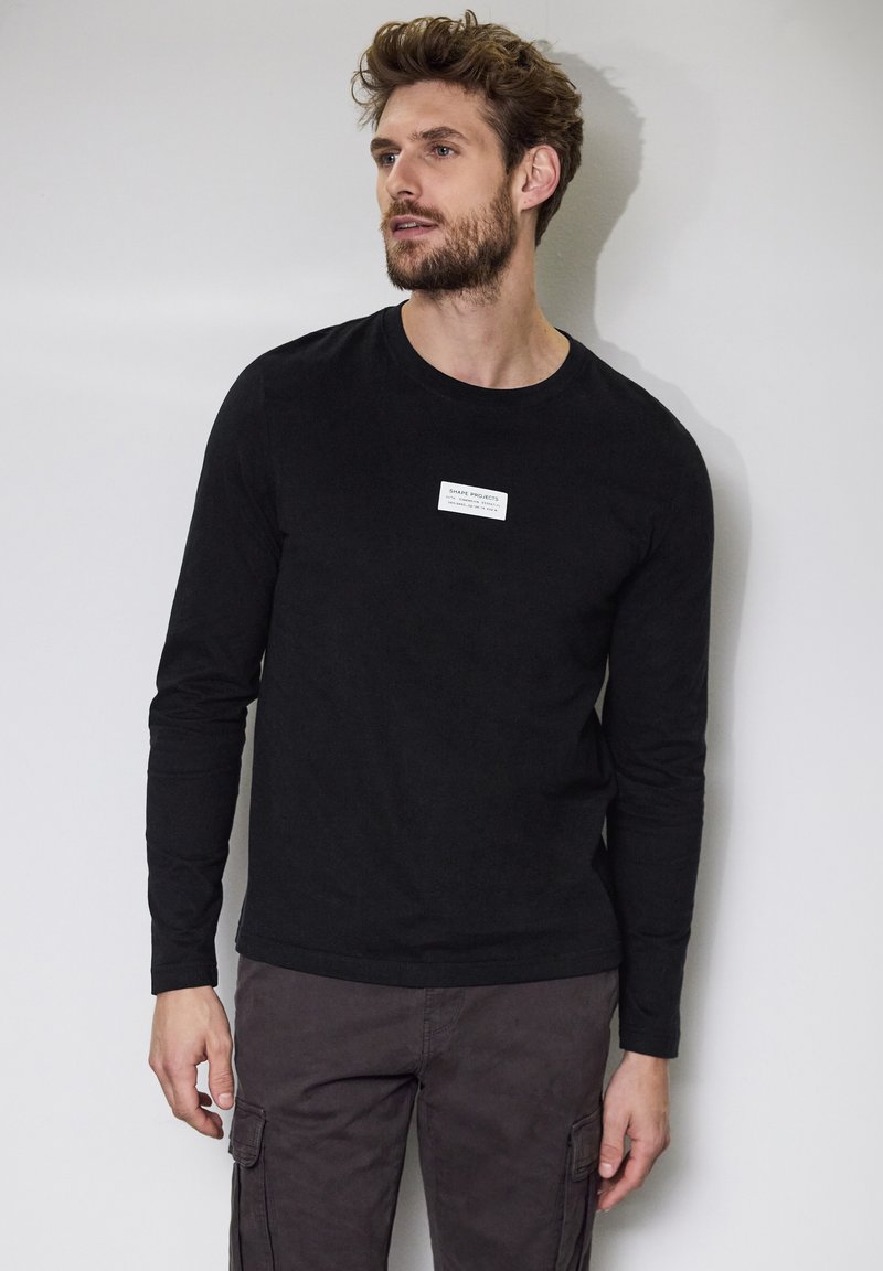 Black long-sleeve shirt with a crew neck and a small white label on the chest, paired with dark cargo pants. Smooth cotton texture.
