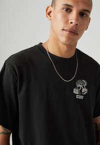 Sort bomulds T-shirt med et grafisk design af et træ, der holder to kufferter, med et lille "Levi's" mærke nær bunden. Klassisk crew neck.
