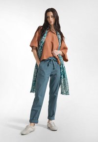 Rostfärgad, ledig topp med korta ärmar, kombinerad med blå byxor med dragsko i midjan och en mönstrad turkos sjal.