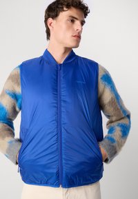 Gilet imbottito blu con una texture liscia e lucida e cerniera frontale, abbinato a un maglione fantasia con dettagli beige e blu.